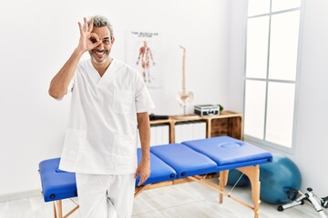 Middle age hispanic therapist man working at pain recovery clinic doing ok gesture with hand smiling, eye looking through fingers with happy face.