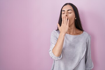 Young brunette woman standing over pink background bored yawning tired covering mouth with hand. restless and sleepiness.