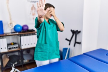 Young hispanic woman wearing physiotherapist uniform standing at clinic covering eyes with hands and doing stop gesture with sad and fear expression. embarrassed and negative concept.