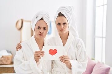 Middle age woman and daughter wearing bath robe holding heart card skeptic and nervous, frowning upset because of problem. negative person.