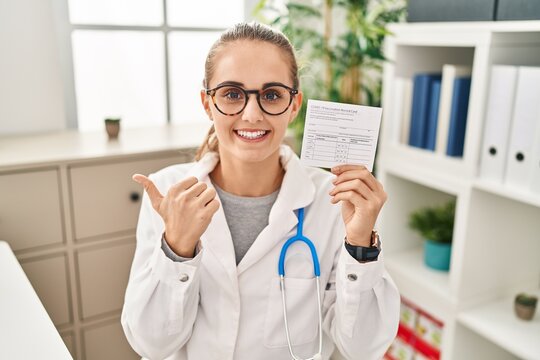 Young doctor woman holding covid certificate pointing thumb up to the side smiling happy with open mouth