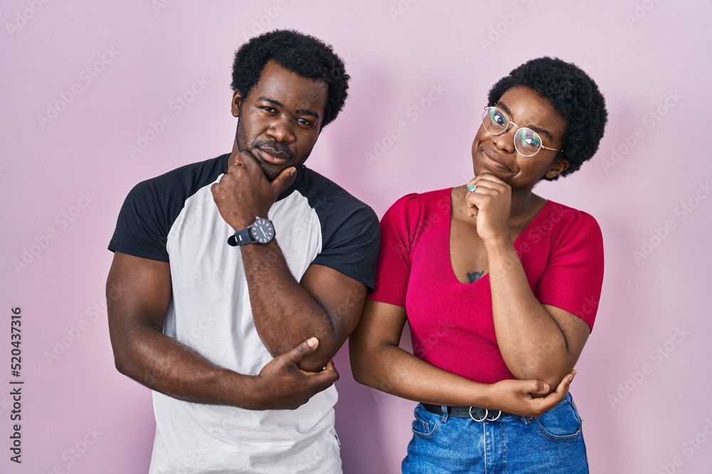 Sticker Young african american couple standing over pink background looking confident at the camera smiling with crossed arms and hand raised on chin. thinking positive.