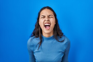 Young brazilian woman standing over blue isolated background angry and mad screaming frustrated and furious, shouting with anger. rage and aggressive concept.