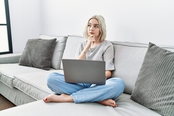 Young caucasian woman using laptop at home sitting on the sofa looking confident at the camera with smile with crossed arms and hand raised on chin. thinking positive.