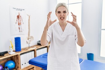 Young caucasian woman working at pain recovery clinic shouting frustrated with rage, hands trying to strangle, yelling mad