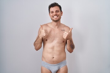 Fototapeta na wymiar Young hispanic man standing shirtless wearing underware success sign doing positive gesture with hand, thumbs up smiling and happy. cheerful expression and winner gesture.