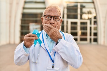 Senior doctor with grey hair holding blue ribbon covering mouth with hand, shocked and afraid for...