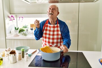 Senior man with grey hair cooking spaghetti at home kitchen screaming proud, celebrating victory and success very excited with raised arms