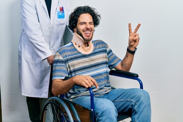 Handsome hispanic man sitting on wheelchair wearing neck collar smiling with happy face winking at...