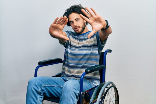 Handsome hispanic man sitting on wheelchair doing frame using hands palms and fingers, camera perspective