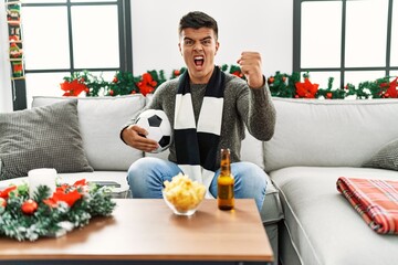 Young hispanic man football hooligan holding ball and beer annoyed and frustrated shouting with anger, yelling crazy with anger and hand raised