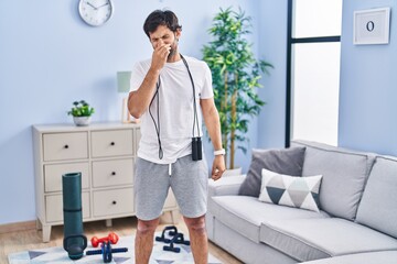 Handsome latin man wearing sportswear at home smelling something stinky and disgusting, intolerable smell, holding breath with fingers on nose. bad smell - obrazy, fototapety, plakaty