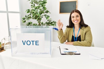 Young brunette woman sitting at election table with voting ballot smiling positive doing ok sign...