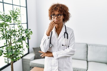 Young african american woman wearing doctor uniform and stethoscope with hand on chin thinking about question, pensive expression. smiling with thoughtful face. doubt concept.