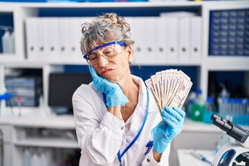 Middle age woman working at scientist laboratory holding dollars serious face thinking about question with hand on chin, thoughtful about confusing idea