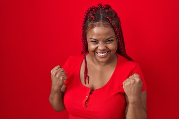 African american woman with braided hair standing over red background very happy and excited doing winner gesture with arms raised, smiling and screaming for success. celebration concept.