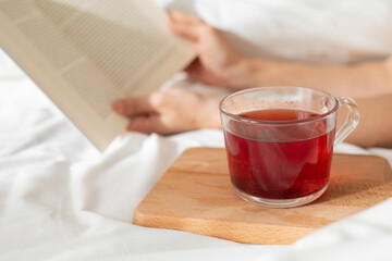 Slow morning concept. Woman's hands, lying on a bed with white sheets reading a book, and with a red tea prepared.