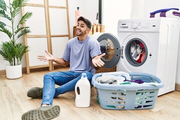 Young hispanic man putting dirty laundry into washing machine smiling cheerful with open arms as friendly welcome, positive and confident greetings