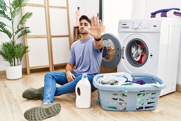 Young hispanic man putting dirty laundry into washing machine doing stop sing with palm of the hand. warning expression with negative and serious gesture on the face.