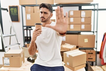 Young hispanic man working at small business ecommerce drinking a coffee with open hand doing stop sign with serious and confident expression, defense gesture