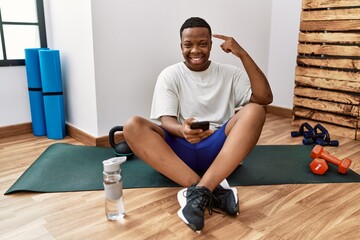 Young african man sitting on training mat at the gym using smartphone smiling pointing to head with one finger, great idea or thought, good memory