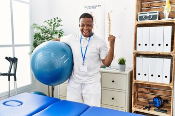 Young african physiotherapy man holding pilates ball at rehabilitation clinic screaming proud,...