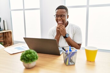 Young african man working at the office using computer laptop laughing nervous and excited with hands on chin looking to the side