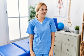 Beautiful blonde physiotherapist woman working at pain recovery clinic looking away to side with smile on face, natural expression. laughing confident.