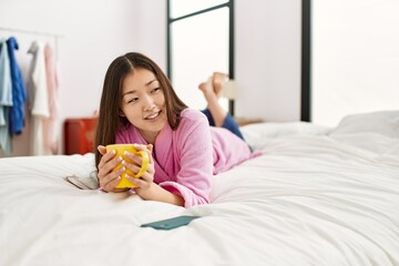 Young chinese girl drinking coffee lying on the bed at bedroom.