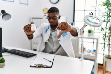 Young african american doctor man wearing doctor uniform holding prescription pills at the clinic with angry face, negative sign showing dislike with thumbs down, rejection concept