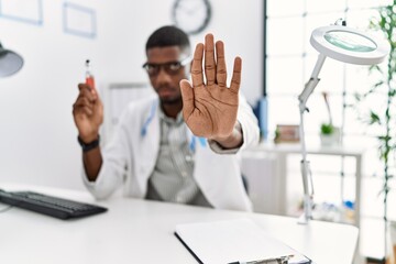 Young african american doctor man holding electronic cigarette at medical clinic with open hand doing stop sign with serious and confident expression, defense gesture