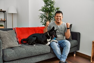 Young latin man and dog sitting on the sofa at home excited for success with arms raised and eyes closed celebrating victory smiling. winner concept.