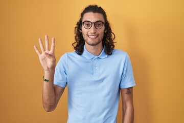 Young hispanic man standing over yellow background showing and pointing up with fingers number four while smiling confident and happy.