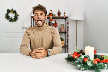 Young handsome man with beard sitting on the table by christmas decoration sticking tongue out happy with funny expression. emotion concept.