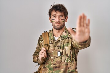 Hispanic young man wearing camouflage army uniform doing stop sing with palm of the hand. warning expression with negative and serious gesture on the face.