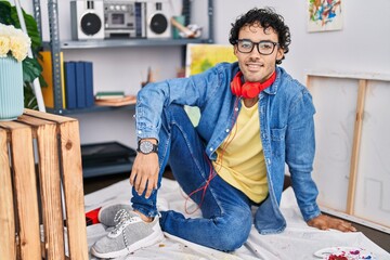 Young hispanic man artist wearing headphones sitting on floor at art studio