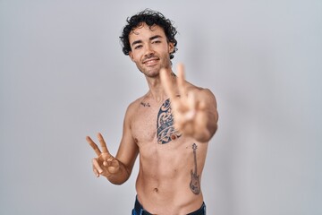 Hispanic man standing shirtless smiling looking to the camera showing fingers doing victory sign. number two.