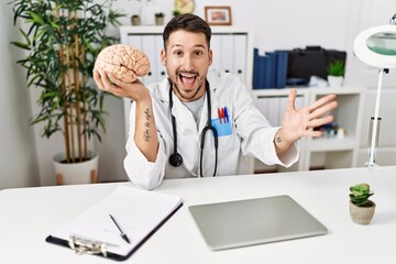 Obraz premium Young doctor holding brain at medical clinic celebrating victory with happy smile and winner expression with raised hands