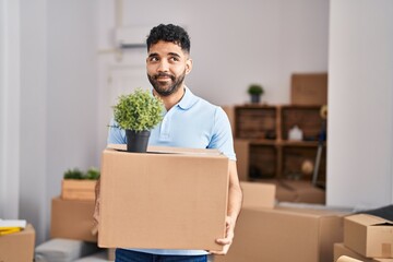 Hispanic man with beard moving to a new home holding box smiling looking to the side and staring away thinking.