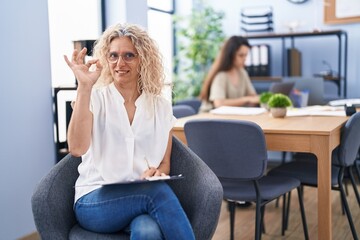 Middle age woman working at the office holding clipboard doing ok sign with fingers, smiling friendly gesturing excellent symbol