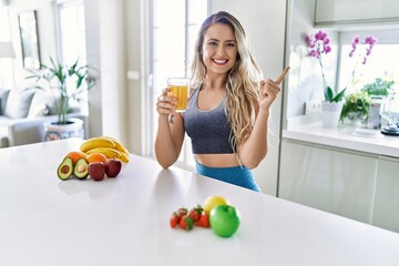 Young caucasian fitness woman wearing sportswear drinking healthy orange juice smiling happy pointing with hand and finger to the side