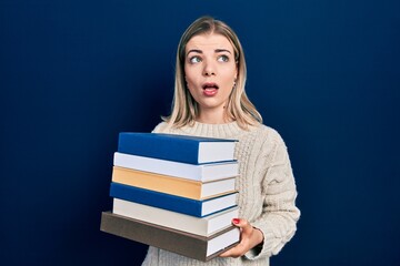 Beautiful caucasian woman holding a pile of books in shock face, looking skeptical and sarcastic, surprised with open mouth