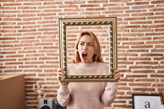 Hispanic woman holding empty frame angry and mad screaming frustrated and furious, shouting with anger. rage and aggressive concept.