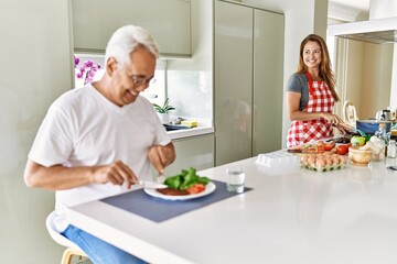 Middle age hispanic man smiling happy eating beef with salad while woman cook at the kitchen.