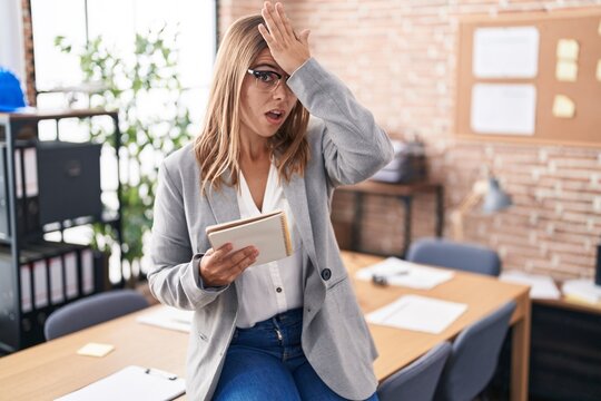 Young hispanic woman working at the office wearing glasses surprised with hand on head for mistake, remember error. forgot, bad memory concept.