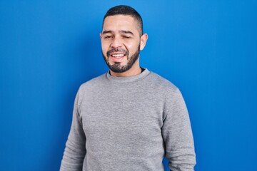 Hispanic man standing over blue background winking looking at the camera with sexy expression, cheerful and happy face.