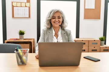 Middle age businesswoman sitting on desk working using laptop at office with a happy and cool smile on face. lucky person.