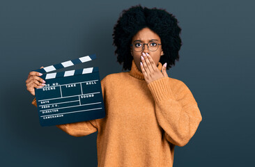 Young african american woman holding video film clapboard covering mouth with hand, shocked and...