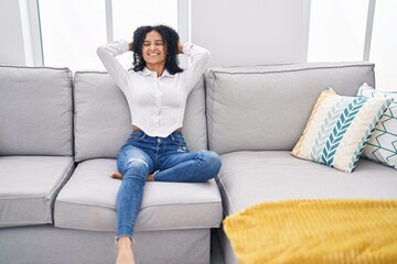 Young hispanic woman relaxed with hands on head sitting on sofa at home