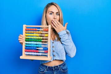 Beautiful caucasian woman with blond hair holding traditional abacus covering mouth with hand, shocked and afraid for mistake. surprised expression